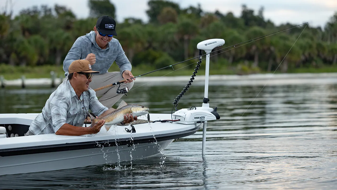 A man holds up a redfish over the edge of their saltwater fishing boat, a trolling motor is visible on the bow.